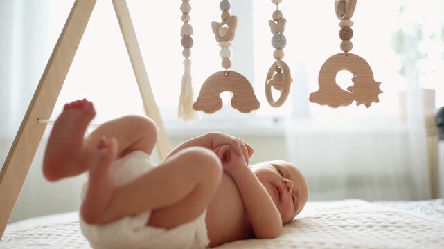 Cute newborn baby boy in a diaper lying on his back on a comfortable bed, actively playing with a stylish wooden baby gym mobile and learning to grasp eco friendly toys