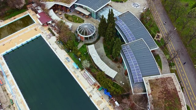 A public swimming pool is seen from above, surrounded by buildings and green trees. Pathways lead through the area, and there are empty chairs and tables waiting for visitors.