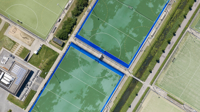 Aerial view of Sportpark De Meerpaal featuring green and blue artificial turf fields, a modern building with solar panels, and a narrow canal in Houten, Utrecht, Netherlands.
