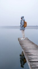 Obraz premium Person in coat and backpack standing on a quiet lakeside dock in soft mist with subtle reflection for minimalist background design
