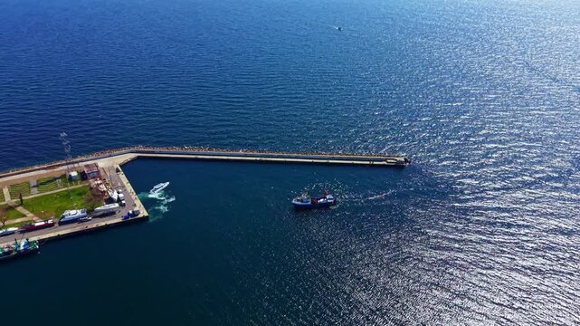 A boat nears the dock along the shoreline. People are seen on the walkway as water sparkles under bright sunlight. Clear blue skies surround the area.