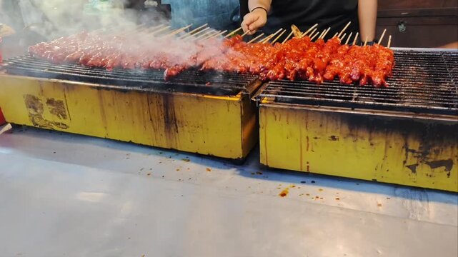 Close-up of street food vendor grilling rows of chicken satay skewers with red sauce on hot charcoal grill with smoke at night market, Asia.