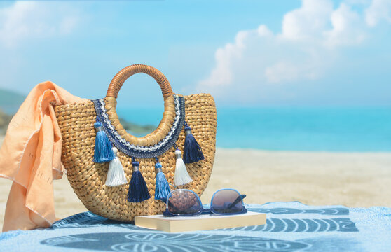 Straw beach bag with tassels, sunglasses and book on towel against blurred sea background. Summer vacation concept