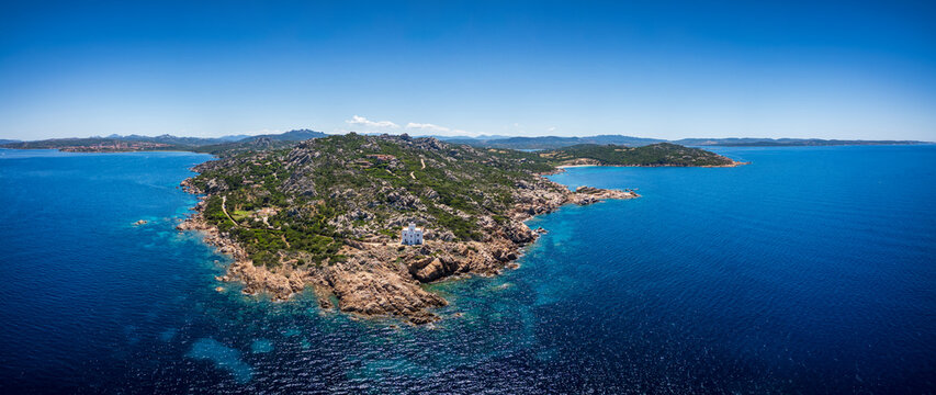 Aerial view of Faro di Punta Sardegna lighthouse on the rocky cliffs of a rugged coastline surrounded by turquoise Mediterranean waters and green vegetation in Palau, Sardegna, Italy.