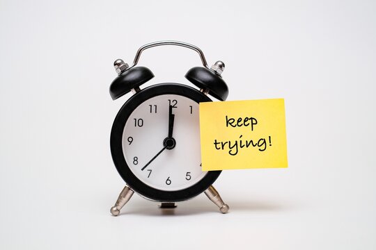 Black alarm clock with motivational sticky note &ldquo;keep trying!&rdquo;. Concept of motivational reminder to maintain persistence and overcome challenges over time