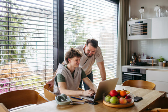 Father helping teenage son with diabetes with homework on laptop in kitchen.