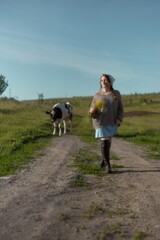 Fototapeta premium Walking down a scenic farm path, a woman enjoys nature with a bouquet in hand as a cow meanders nearby