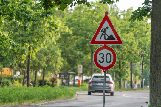 A roadworks warning sign stands above a thirty speed limit sign. Green deciduous trees frame a calm residential street.