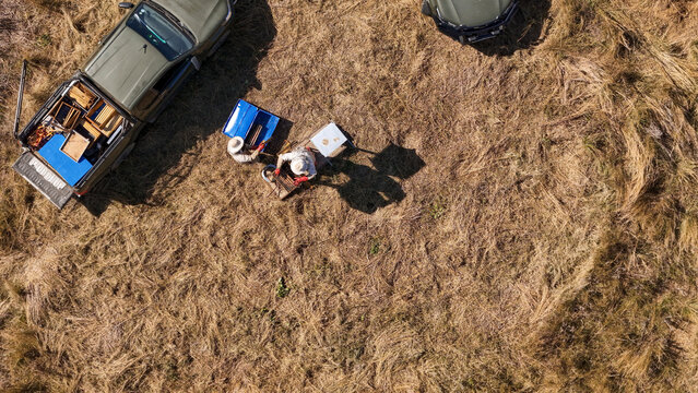 Aerial view of beekeepers in protective suits working with beehives and honey frames next to a pickup truck in a dry field in Pretoria, Gauteng, South Africa.