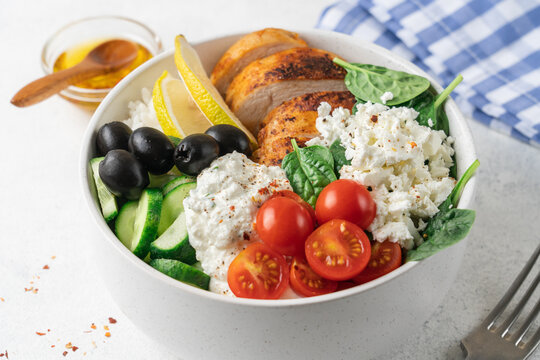 Healthy high protein Mediterranean chicken bowl with rice, cucumber, cherry tomatoes, feta cheese, olives, spinach and creamy yogurt sauce close up view on light background with copy space