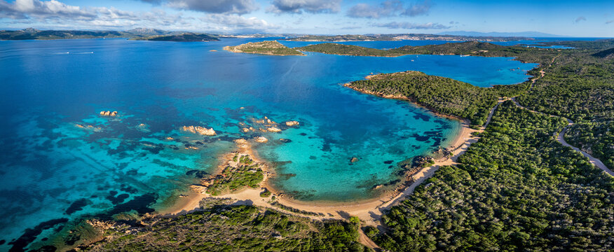 Aerial view of Spiaggia di Porto Palma with its turquoise waters, sandy crescent beach, and rugged green coastline under a bright sky La Maddalena, Sardegna, Italy.