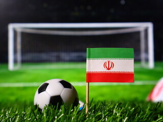 Small flag of iran on a wooden stick stuck in the green grass of a soccer field with a soccer ball and a goal post in the background © Photo Agency
