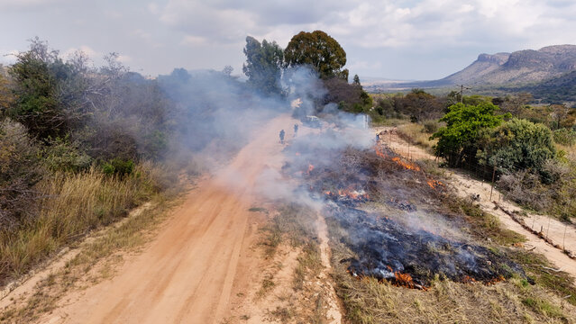 Aerial view of a controlled bushfire burning along a dirt road with smoke rising and workers monitoring the flames near mountains in Vaalwater, Limpopo, South Africa.