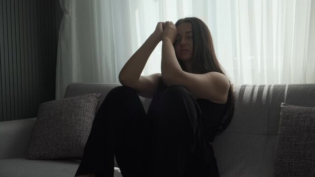 Young woman sitting on sofa near window in deep emotional distress, alone in silence facing inner struggles and mental health challenges