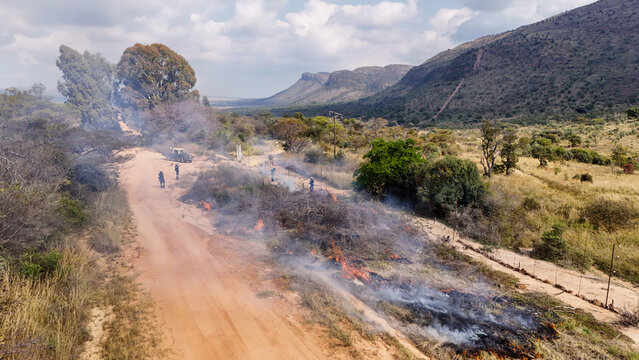 Aerial view of a controlled brush fire along a dirt road with workers managing the flames in a rural landscape with mountains in Vaalwater, Limpopo, South Africa.