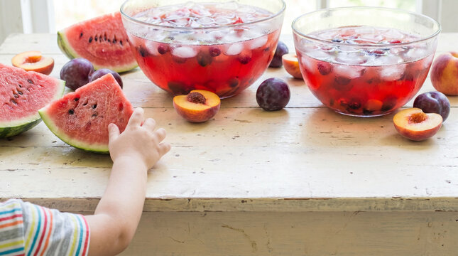 Child reaching for a slice of watermelon on a rustic white wooden table. Summer party setup with fresh fruit and large glass bowls of red iced punch. Copy space for text