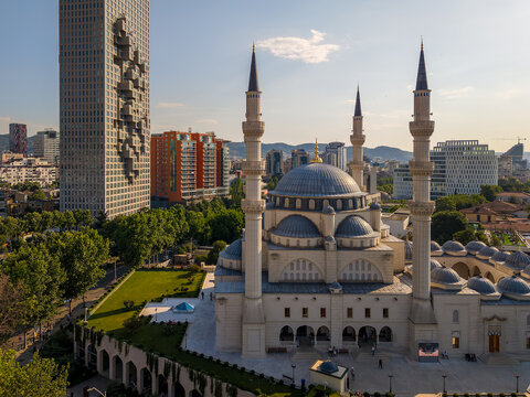 Aerial view of the Mosque of Namazgah with its grand domes and minarets next to a modern pixelated skyscraper and green trees in Tirane, Tirana County, Albania.