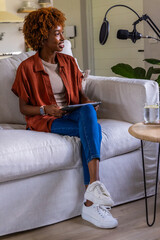 Obraz premium African American woman sitting on sofa wearing rust shirt holding tablet speaking into mic at home