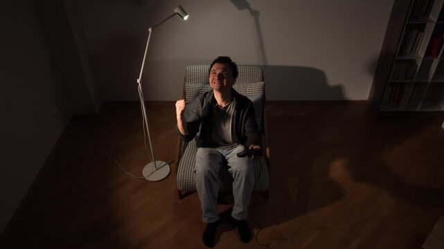Happy man sitting in a comfortable chair, holding a controller, expressing joy and excitement while watching tv or playing video games in a dimly lit room with a floor lamp