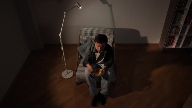 Man sitting on an armchair in a dimly lit living room, relaxing and consuming potato chips from a bowl, enjoying a casual evening snack under a floor lamp