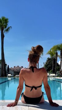 backview of woman in black bikini sitting by luxury resort swimming pool, authentic summer vacation lifestyle