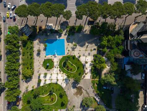 Aerial view of a public square featuring a blue sports court, lush green lawns, and tree-lined streets in Tirane, Tirana County, Albania.