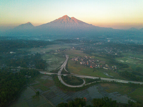 Aerial view of a modern highway interchange surrounded by lush rice fields and a traditional village with a large volcano in the background in Indonesia.