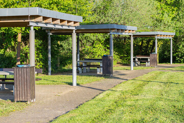 Fototapeta premium Outdoor picnic area featuring wooden tables under shaded pergolas surrounded by lush greenery and well-maintained grass in a public park setting