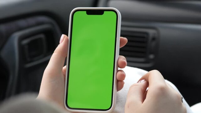 Hands hold phone with green screen inside car. Fingers tap screen while seated. Suitable for app demos, navigation, or mobile usage in vehicle. Focus on phone and interaction