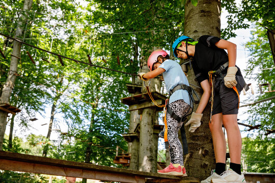 Teenager Boy helping girl on rope course in forest adventure park, children wearing helmets and harnesses supporting each other, outdoor activity. Brother and little sister, siblings, having fun