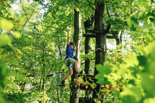 Teenage boy climbing rope course in forest adventure park, child wearing helmet and harness enjoying outdoor challenge, real life.