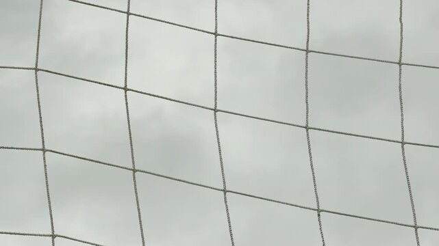 A wet soccer net hangs quietly from a weathered metal crossbar, creating a simple grid pattern. The gloomy, overcast sky brings a feeling of calm and quiet solitude to the empty field.