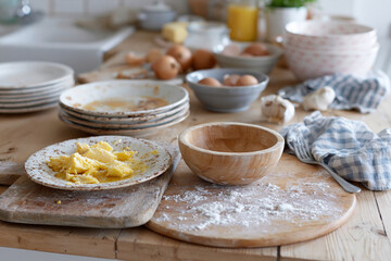 Fototapeta premium Cozy kitchen scene with scattered cooking tools and ingredients. Flour-dusted board holds a wooden bowl and yellow butter. Stacked plates, eggs, and a checkered towel add lived-in charm