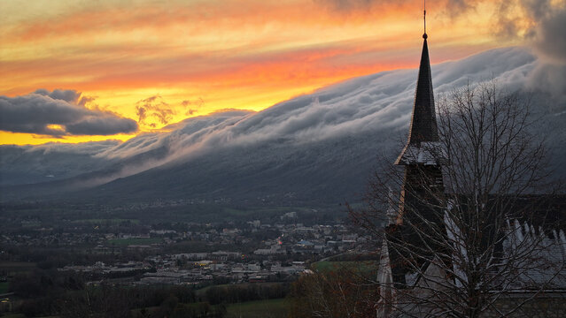 Aerial view of Chapelle Notre-Dame de Riantmont with its sharp spire against a dramatic sunset and clouds rolling over the Jura mountains in Vesancy, Auvergne-Rhone-Alpes, France.