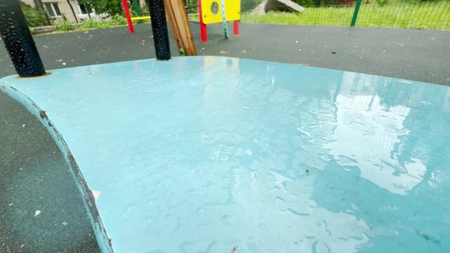a wet blue swing seat moves in the wind. The motion creates a rhythmic, metallic creaking sound. Water droplets splash softly onto the ground below.