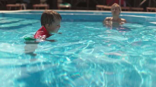 Boy in oversleeves swims in a pool of clear water with mom and sister on summer vacation