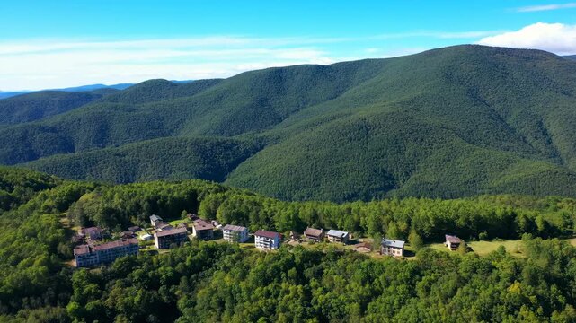 Aerial view of a small mountain village surrounded by dense forest and green ridges in clear daylight