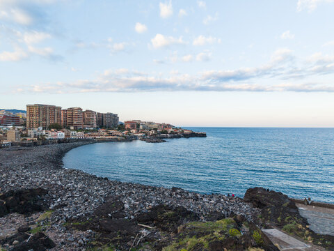 Aerial view of the volcanic rocky coastline and urban buildings along the Mediterranean Sea under a clear sky in Catania, Sicily, Italy.