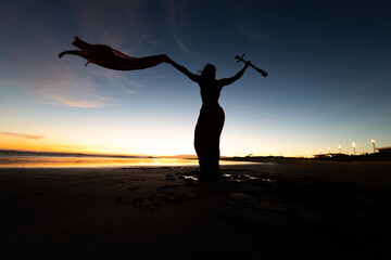 Fototapeta premium Silhouette of a woman in a flowing dress holding a stick against a colorful sunset at the beach, with reflections on the wet sand and distant lights