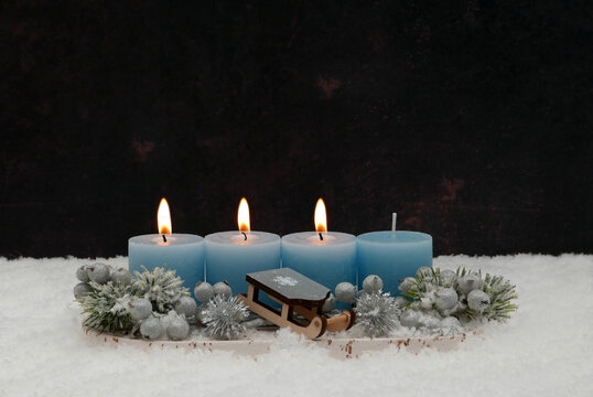 Third Sunday of Advent: An arrangement featuring three burning blue candles, a small wooden sled, and fir branches in the snow, set against a dark background with ample space for text.