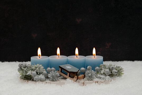 Fourth Sunday of Advent: An arrangement featuring four burning blue candles, a small wooden sled, and fir branches in the snow, set against a dark background with ample space for text.