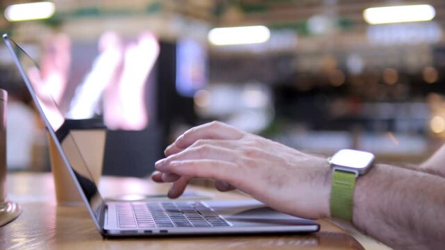 Hands type on laptop in cozy cafe setting. Person wears green watch and focuses intently. Background shows warm, blurred lights and cafe ambiance. Ideal for remote work, freelancing