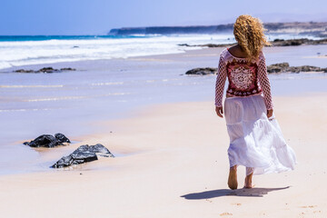 Woman walking on a sandy beach in a white flowing skirt and crochet top, strolling along the ocean shoreline for a peaceful sunny seaside summer escape © simona