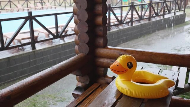 An inflatable duck lies on wooden table in gazebo at recreation center against the backdrop of pool covered with rain