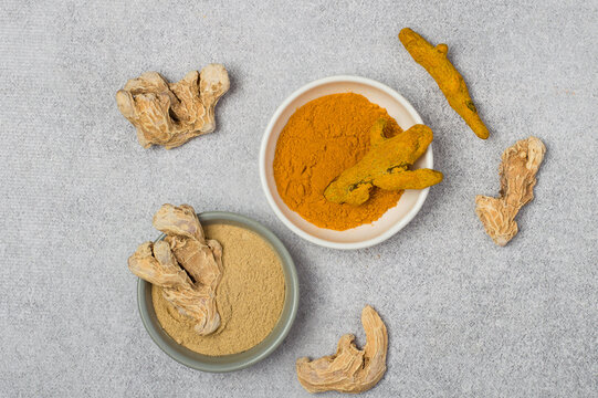 Organic ground ginger and turmeric powder with dried roots in ceramic bowls, on grey background.