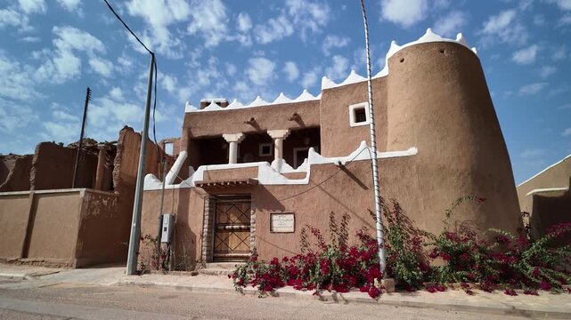 Ushaiqer Heritage Village preserving Najdi mud-brick architecture with white decorative trim, round tower and vivid red bougainvillea against a partly cloudy sky, historic charm of Saudi Arabia