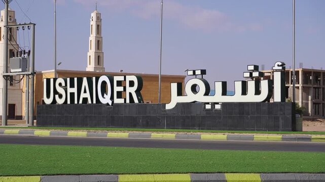 Ushaiqer village entrance sign in both English and Arabic, visible by the road with a mosque minaret and modern construction in the background under a clear sky of Saudi Arabia
