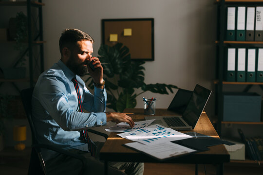 Professional executive reviewing business documents and speaking on phone while working late in office