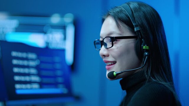 Asian female customer service agent wearing a headset in a modern high-tech office. Smiling technical support operator assisting clients online. Telemarketing consultant working in a dark server room