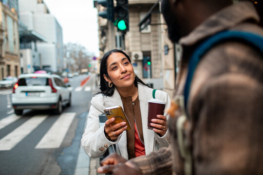 Young woman with coffee and smartphone talking to friend on city street